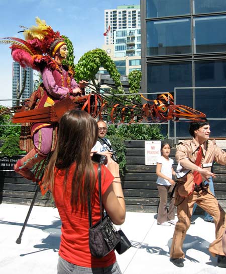Woman riding a wireframe ostrich with oddly-dressed assistant